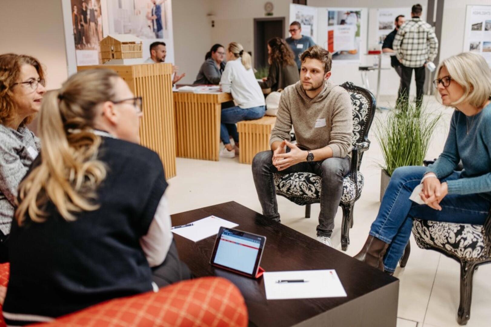 Eine Gruppe von vier Personen sitzt bei einer modernen Indoor-Veranstaltung im Kreis und unterhält sich, während auf dem Tisch Notizblöcke und ein Tablet liegen. Andere Gruppen unterhalten sich im Hintergrund inmitten von Informationsdisplays.