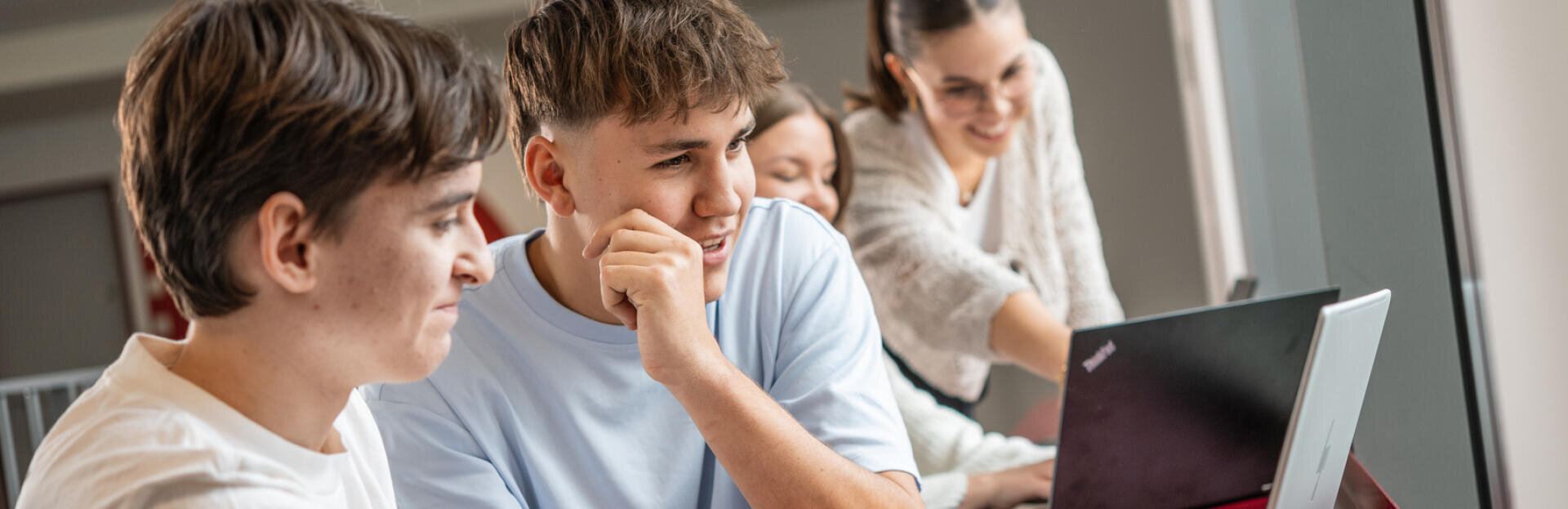 Zwei Jungen im Teenageralter sitzen an einem Schreibtisch, schauen auf einen Laptop und lächeln, während zwei junge Frauen im Hintergrund ebenfalls auf den Laptop schauen, wobei eine von ihnen auf den Bildschirm zeigt, was auf gemeinsames Lernen hindeutet.