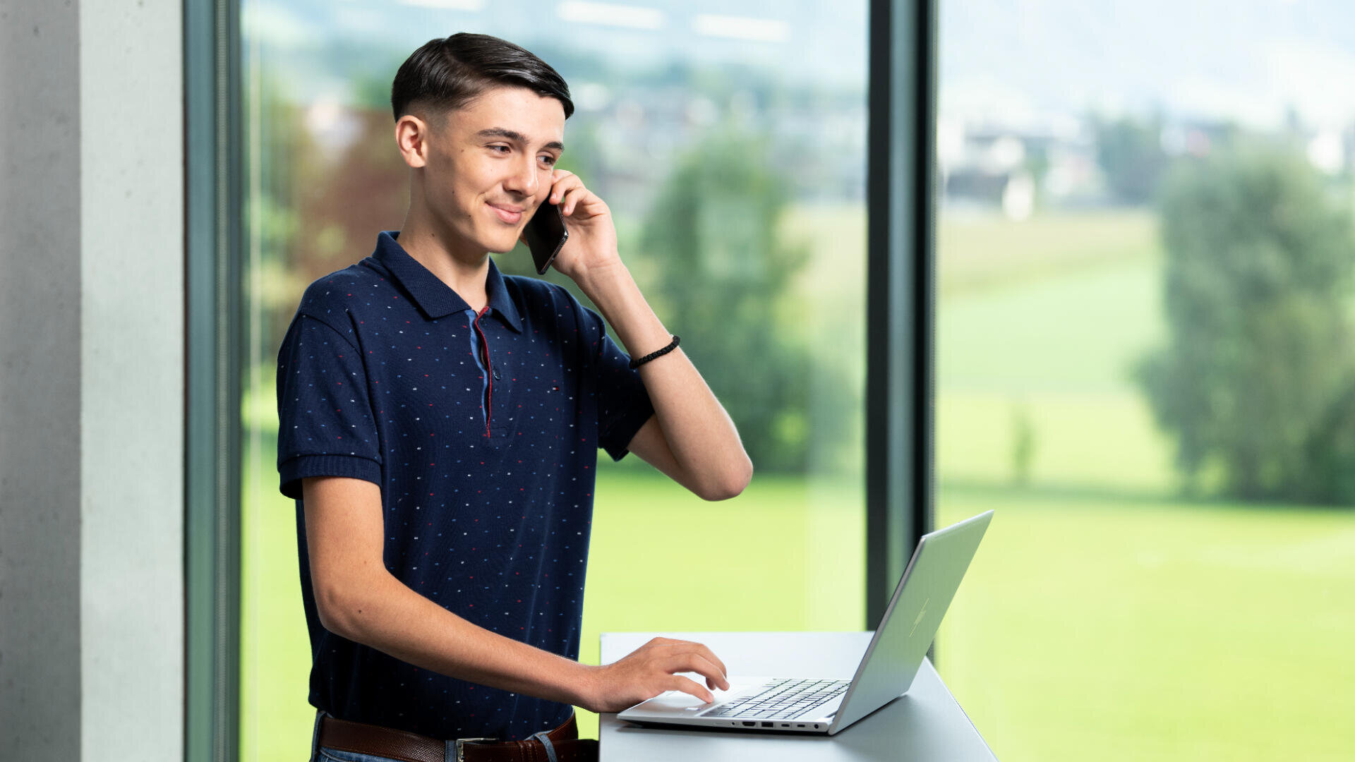 Eine junge Person steht an einem Schreibtisch und spricht mit einem Smartphone, während sie einen Laptop benutzt. Große Fenster hinter ihnen zeigen eine grüne Landschaft mit Bäumen und Hügeln.