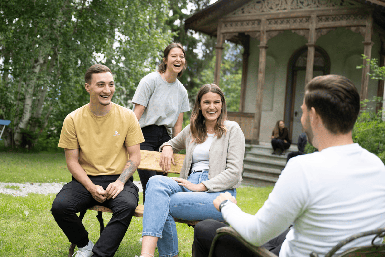 Vier junge Erwachsene sitzen und lachen gemeinsam im Freien auf einer Wiese, mit Bäumen und einem rustikalen Holzhaus im Hintergrund. Eine Person sitzt auf den Stufen, während die Gruppe davor zu plaudern scheint und die Gesellschaft der anderen genießt.