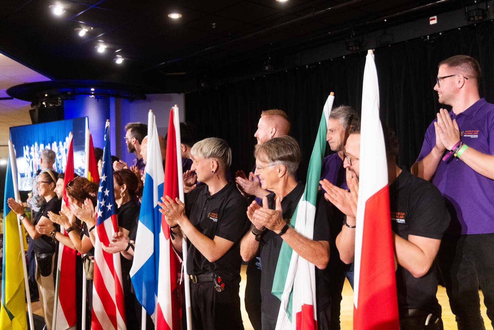 Eine Gruppe von Menschen steht auf der Bühne, hält und klatscht neben verschiedenen Nationalflaggen, darunter die der USA, Frankreichs, Italiens und Polens, während das Publikum auf einem Bildschirm im Hintergrund zu sehen ist.
