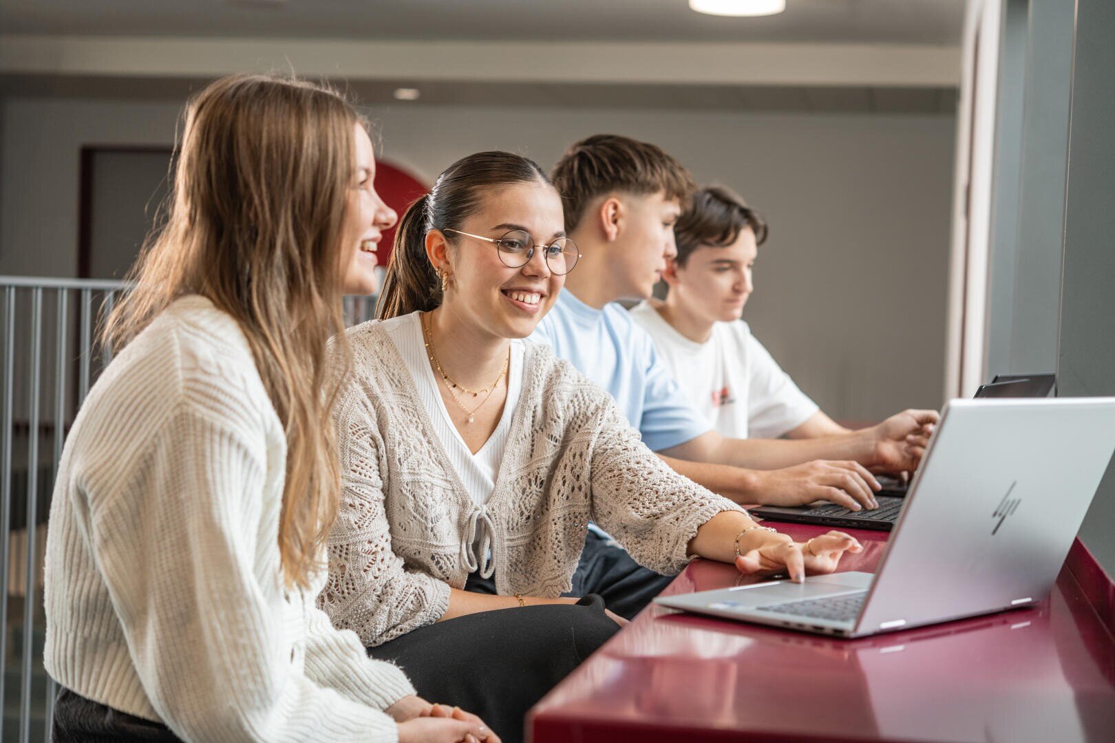Vier junge Erwachsene sitzen drinnen an einem roten Tisch, arbeiten an Laptops und lächeln, während sie sich unterhalten. Zwei Personen blicken in die Kamera, zwei sind auf ihre Bildschirme konzentriert.