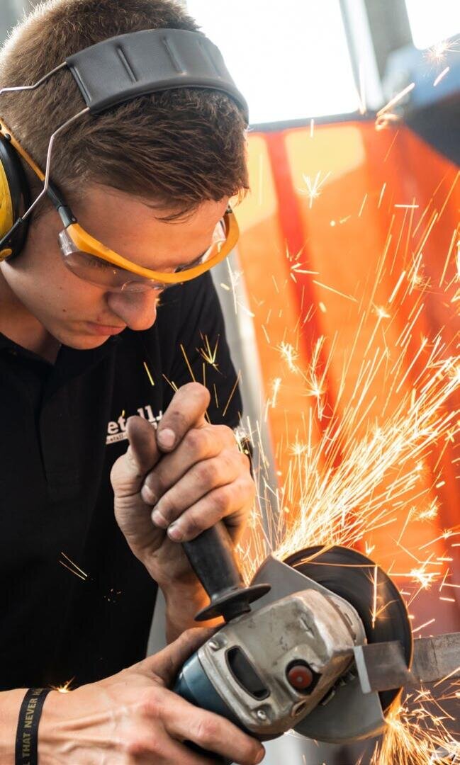 Eine Person, die eine Schutzbrille und einen Gehörschutz trägt, bearbeitet mit einem Winkelschleifer Metall und erzeugt dabei helle Funken.