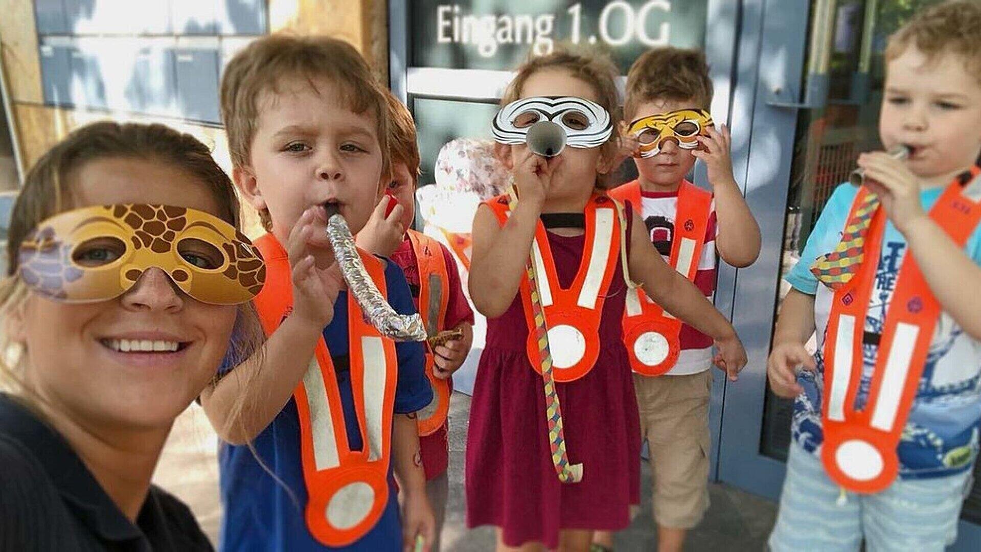 Eine Gruppe junger Kinder und ein Erwachsener tragen Tiermasken und orangefarbene Westen, blasen in Partyblasen und lächeln vor einem Gebäude mit einer Glastür mit der Aufschrift Eingang 1.OG.