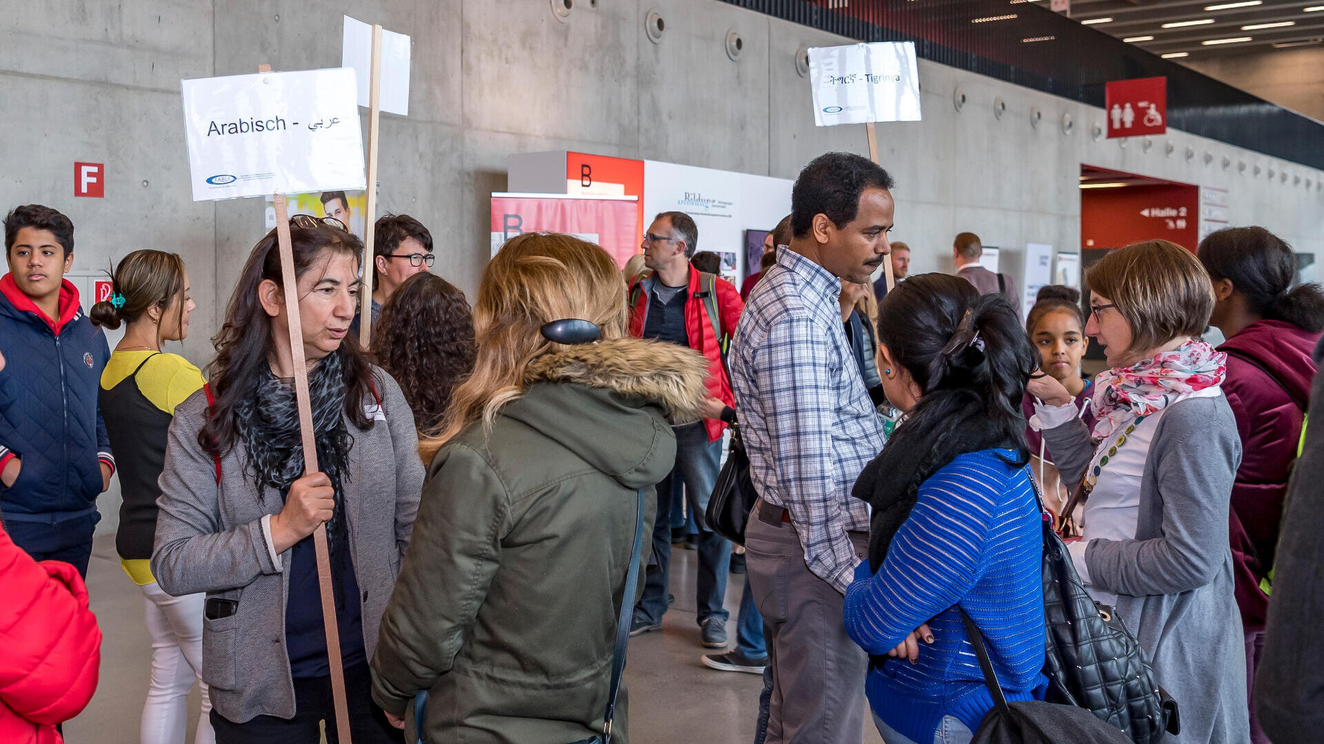 Eine Gruppe von Menschen in einem Innenraum bei einer Veranstaltung, einige halten Schilder mit Namen in verschiedenen Sprachen. Sie unterhalten sich vor einer grauen Betonwand mit Schildern und Buchstaben.