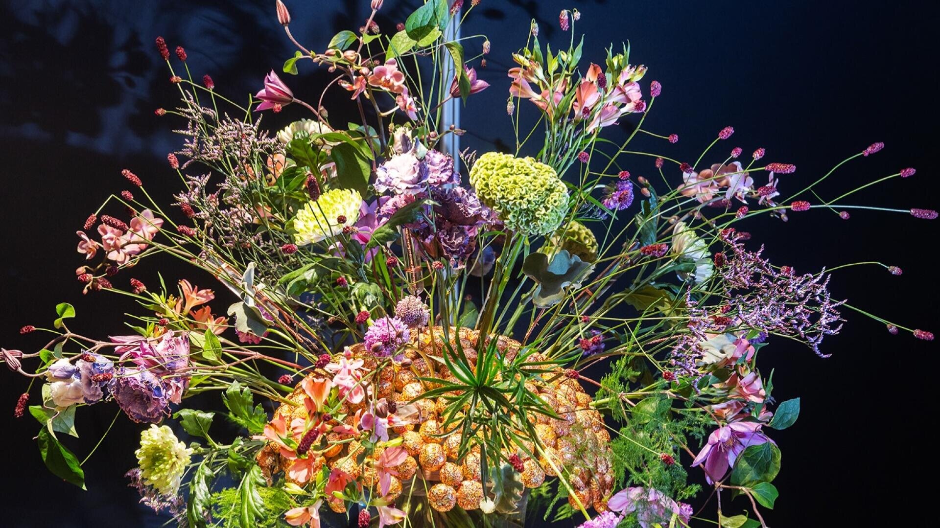 Ein lebendiger, kunstvoller Strauß aus verschiedenen bunten Blumen und Grünpflanzen vor einem dunklen Hintergrund, mit verschlungenen Stängeln und Blüten, die in alle Richtungen ausstrahlen.