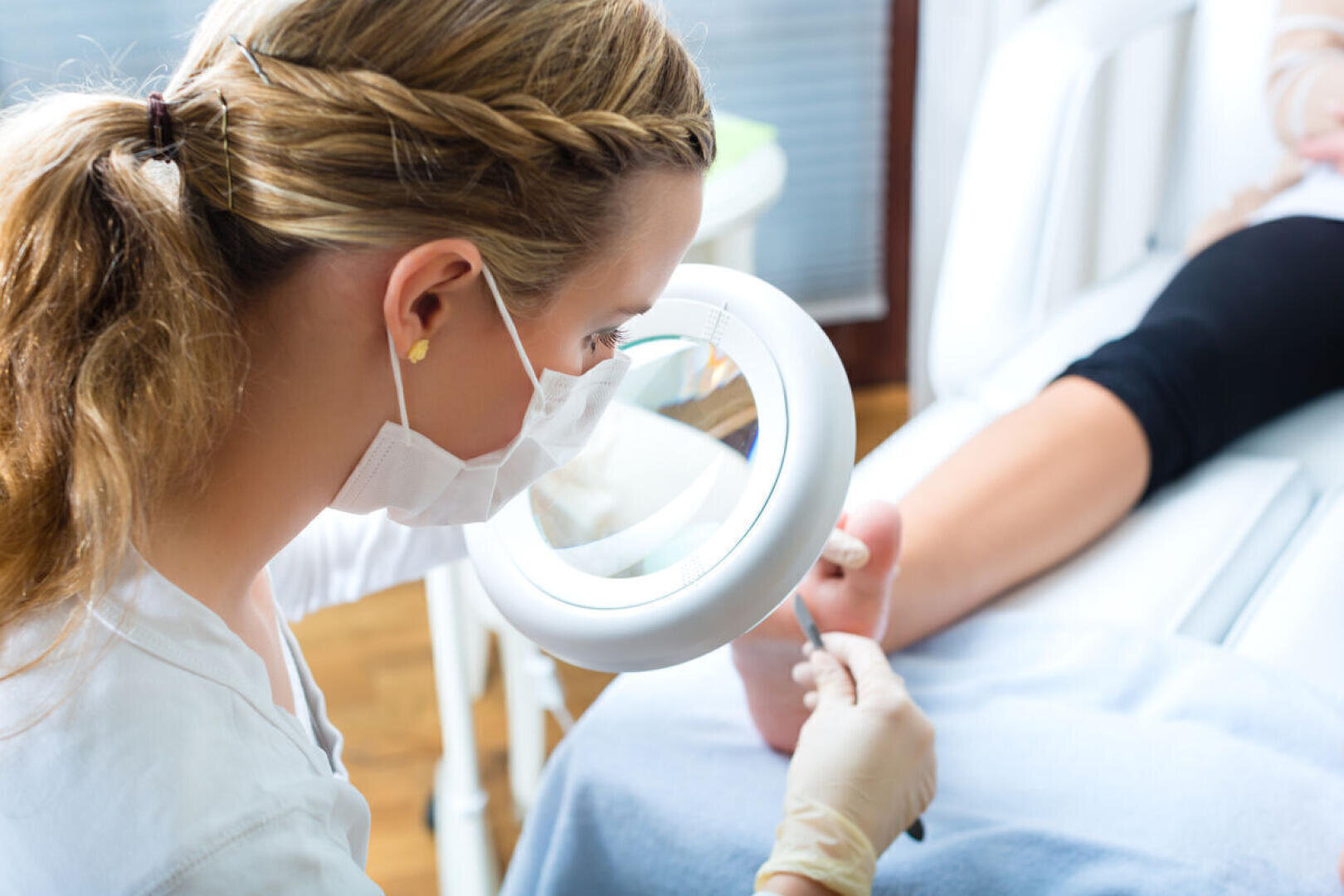 Ein Podologe, der eine Gesichtsmaske und Handschuhe trägt, untersucht den Fuß eines Patienten mit einer Lupenlampe und einem Instrument.