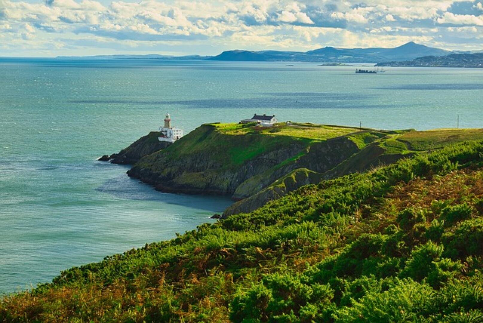 Eine malerische Küstenlandschaft mit einem Leuchtturm und Gebäuden auf einer grasbewachsenen Klippe mit Blick auf ein ruhiges blaues Meer, mit entfernten Bergen und teilweise bewölktem Himmel im Hintergrund.