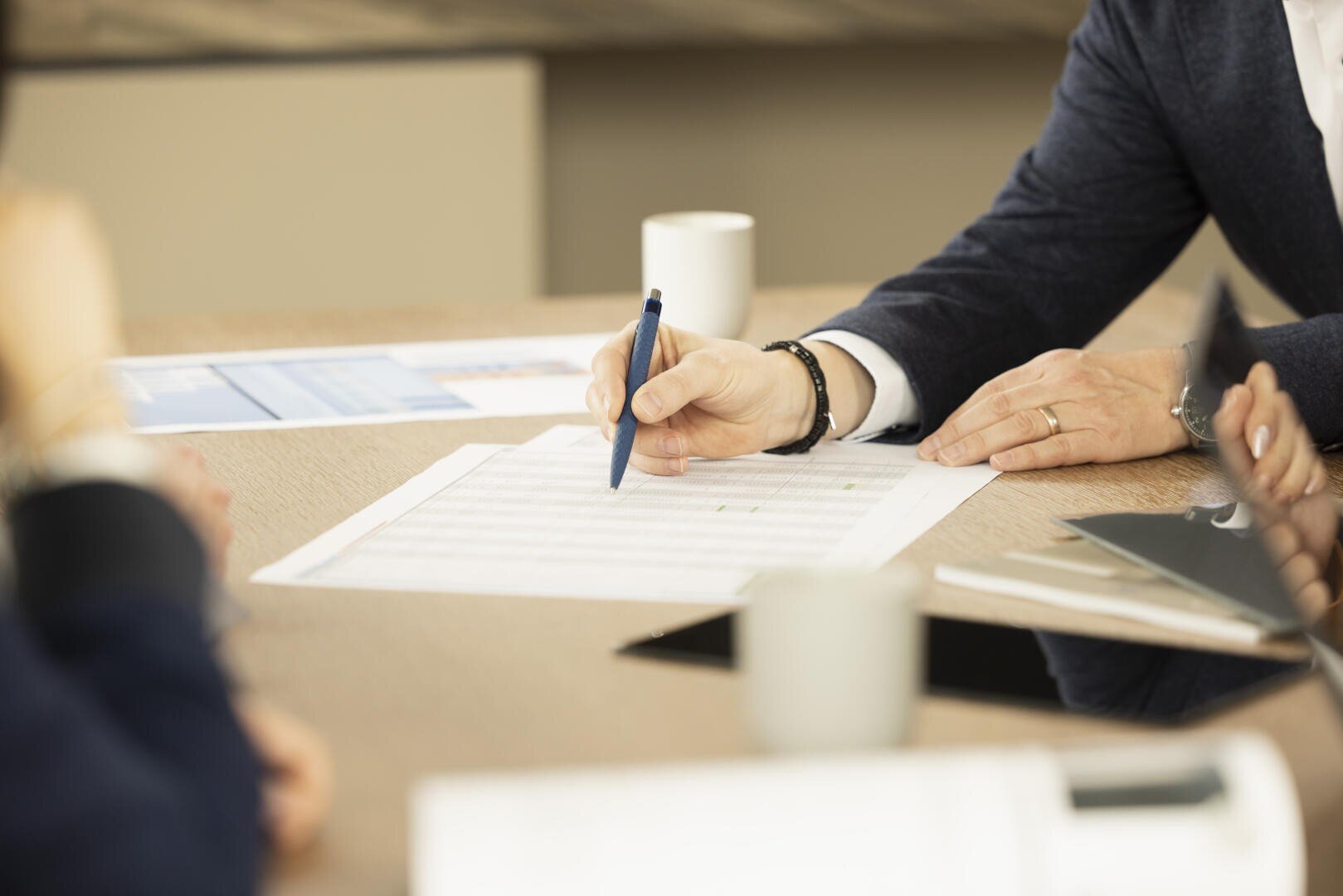 Eine Person in Geschäftskleidung zeigt mit einem blauen Stift auf eine ausgedruckte Tabelle, die auf einem Holztisch liegt. Daneben stehen Kaffeetassen und liegen Unterlagen, und im Hintergrund sind weitere Personen teilweise zu sehen.
