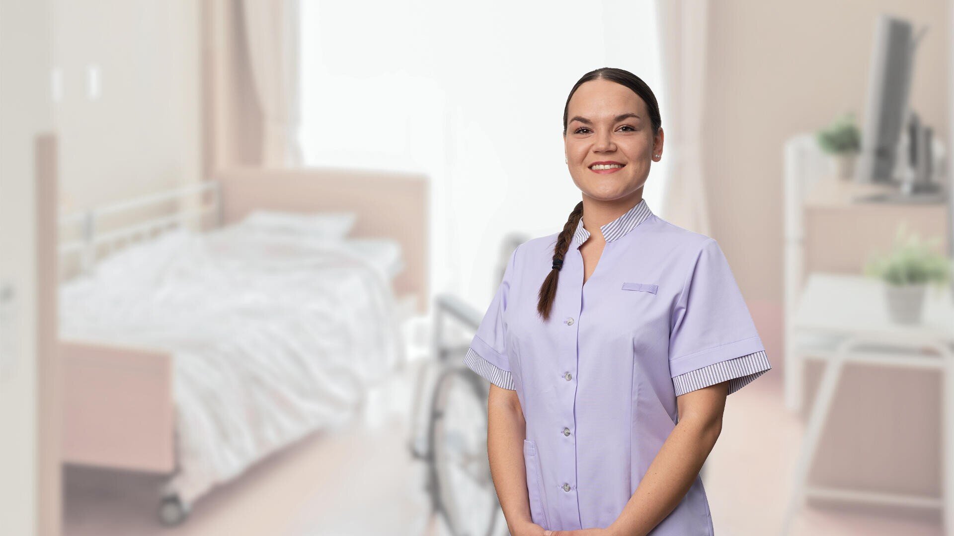 Eine lächelnde Frau in einer hellvioletten Uniform steht in einem hellen, aufgeräumten Zimmer mit einem Bett und einem Rollstuhl im Hintergrund.