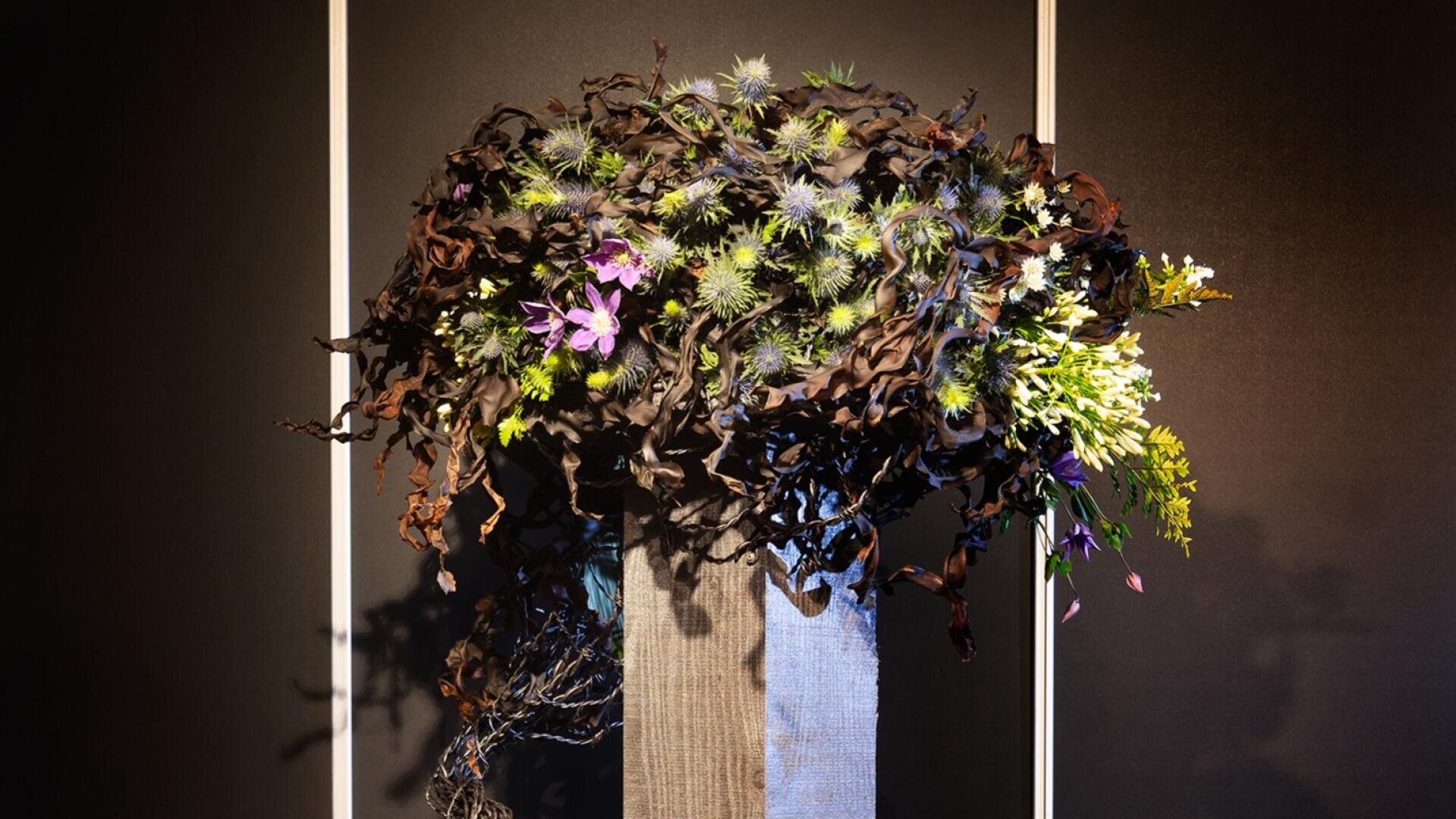 Eine hohe, rechteckige Vase enthält ein kunstvolles Arrangement aus dunklem, gekräuseltem Laub, stacheligen blauen Disteln und kleinen violetten Blüten vor einem dunklen Hintergrund mit zwei vertikalen weißen Linien.