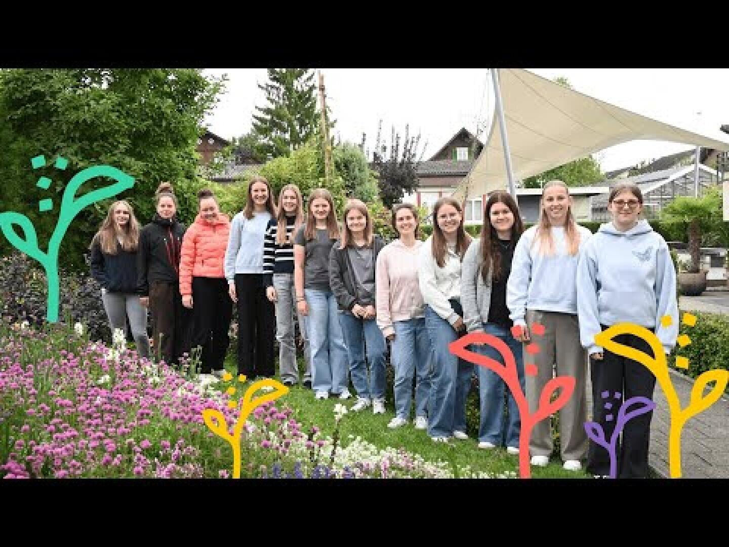 Eine Gruppe von zwölf Frauen steht zusammen in einem Garten mit blühenden Blumen und Grünzeug. Auf dem Foto sind bunte Blumenillustrationen gezeichnet, und im Hintergrund sind Gebäude unter einem bewölkten Himmel zu sehen.