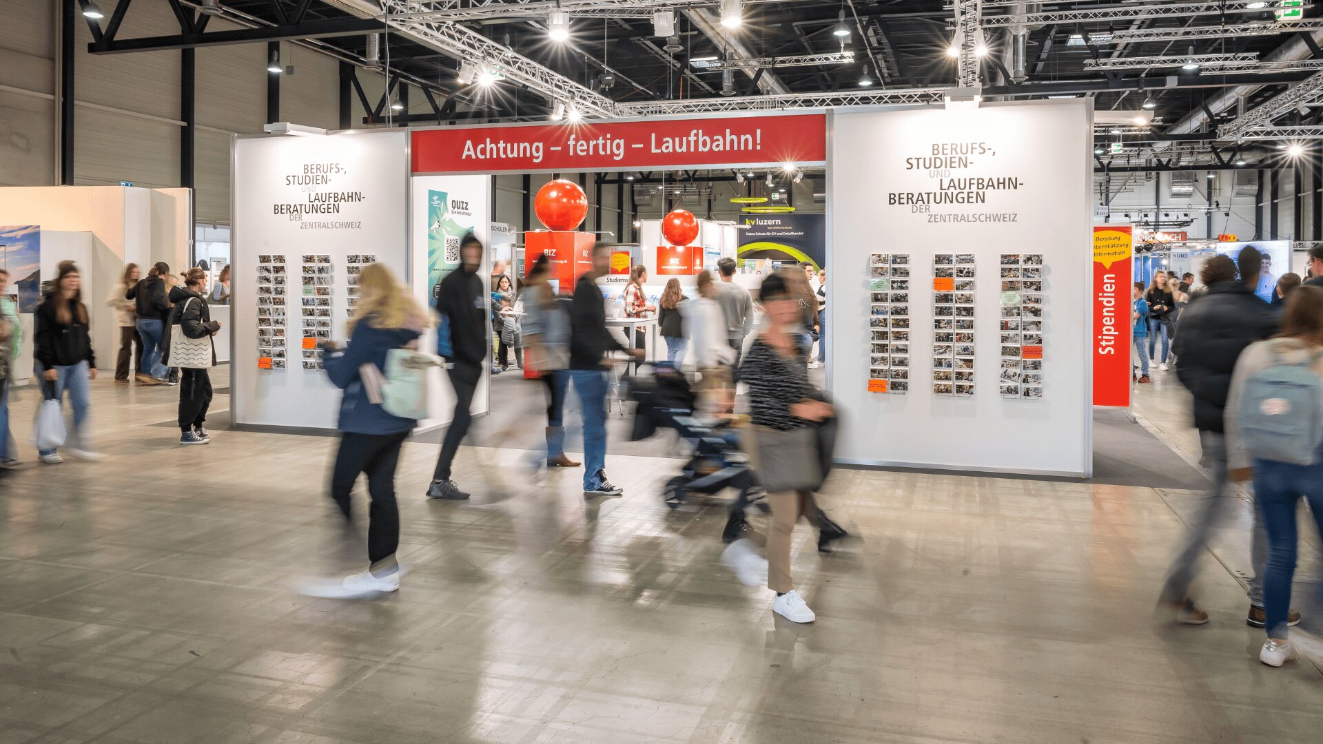 Man geht an einem belebten Stand der Berufs- und Bildungsmesse vorbei, auf dem ein rotes Banner mit der Aufschrift "Achtung - fertig - Laufbahn!" und Informationsdisplays zu sehen sind. Die Kulisse ist eine große, gut beleuchtete Messehalle.