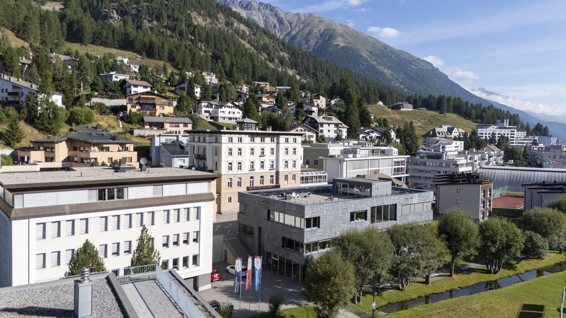 Von Bäumen umgebene Campusgebäude der HFT Graubünden im Engadin, mit Häusern an einem Hang und Bergen im Hintergrund unter blauem Himmel.