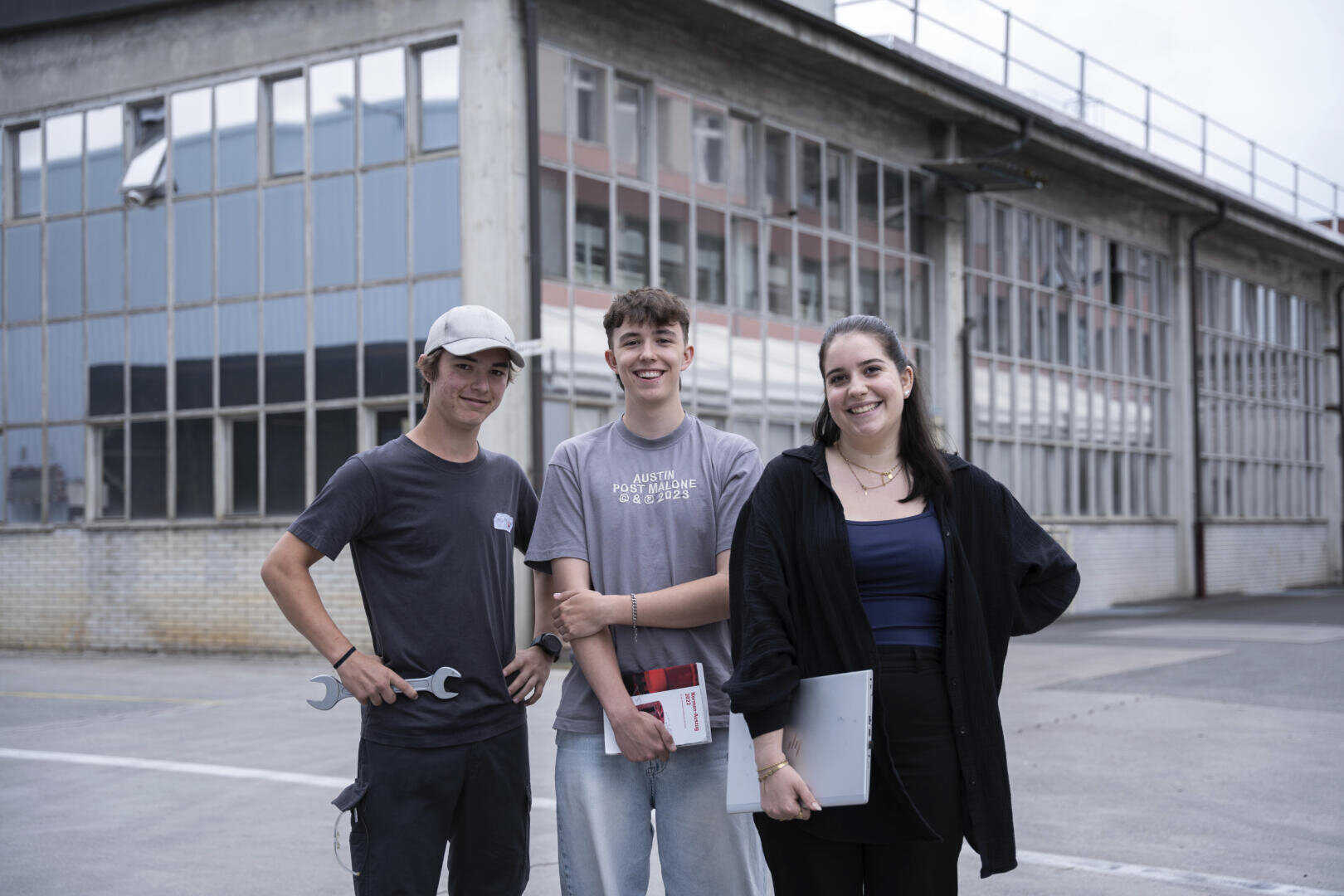 Drei junge Leute stehen vor einem großen Industriegebäude; einer hält einen Schraubenschlüssel in der Hand, ein anderer ein Buch und der dritte einen Laptop.