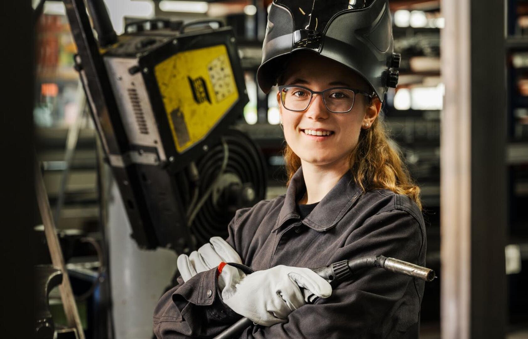 Eine Frau mit Schutzbrille, Schweißerhandschuhen und einem dunklen Schweißerhelm steht in einer Industriehalle, lächelt und hält mit verschränkten Armen einen Schweißbrenner.