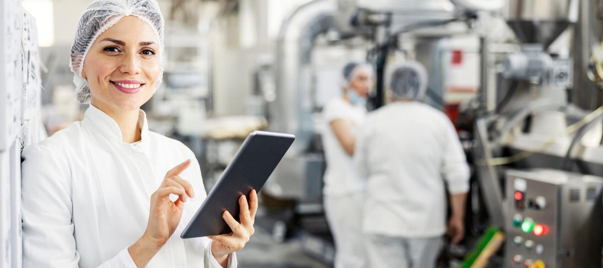 Eine Frau in einem weißen Laborkittel und einer Haube lächelt, während sie in einer Lebensmittelverarbeitungsanlage ein Tablet bedient. Im Hintergrund sind weitere Mitarbeiter in ähnlicher Kleidung zu sehen, die an Maschinen arbeiten.