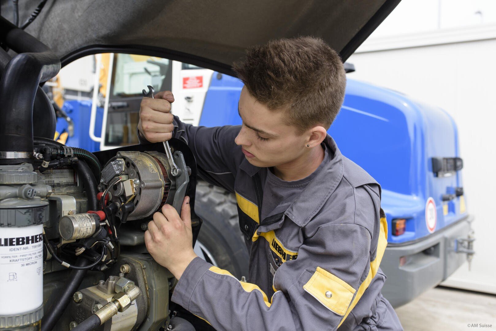 Ein junger Mechaniker in einer grau-gelben Uniform repariert mit einem Schraubenschlüssel Maschinen unter der Motorhaube eines Fahrzeugs, wobei im Hintergrund Teile und ein blauer Lastwagen zu sehen sind.