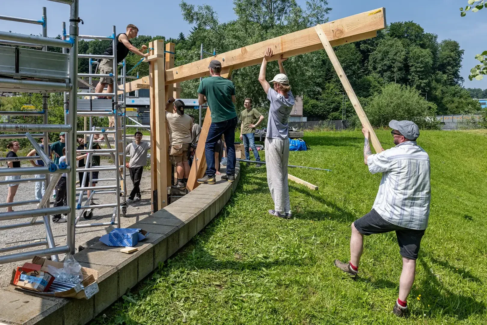 Eine Gruppe von Menschen arbeitet im Freien zusammen, um einen großen Holzbalken mit Hilfe von Gerüsten und Stützen auf einer Rasenfläche neben einer Ziegelmauer zu heben und zu montieren, mit Bäumen und Grünpflanzen im Hintergrund.