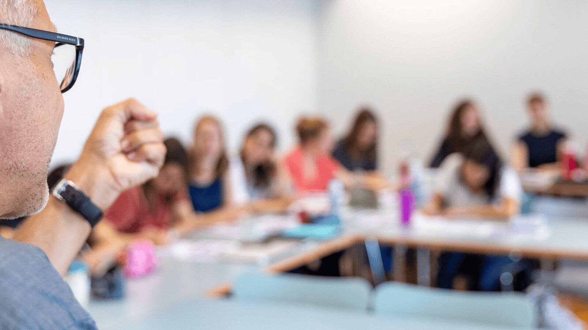 Eine Person gestikuliert, während sie zu einer Gruppe von Schülern spricht, die an Tischen in einem Klassenzimmer sitzen. Der Fokus liegt auf dem Sprecher, während die Schüler und der Hintergrund verschwommen sind.