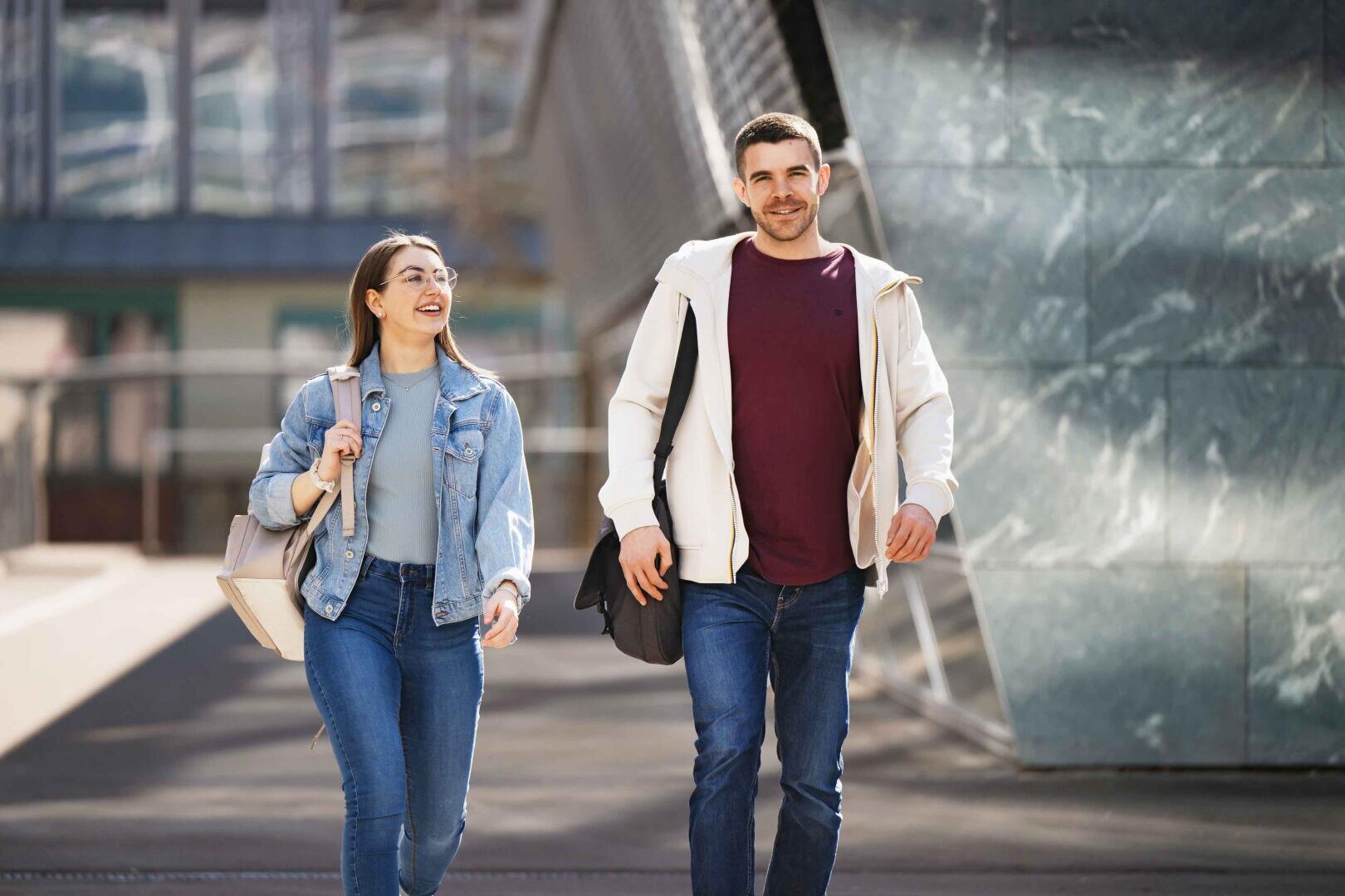 Eine junge Frau und ein junger Mann gehen lächelnd im Freien spazieren, jeder trägt eine Umhängetasche. Sie sind leger in Jeans und Jacken gekleidet, im Hintergrund sind moderne Gebäude zu sehen.