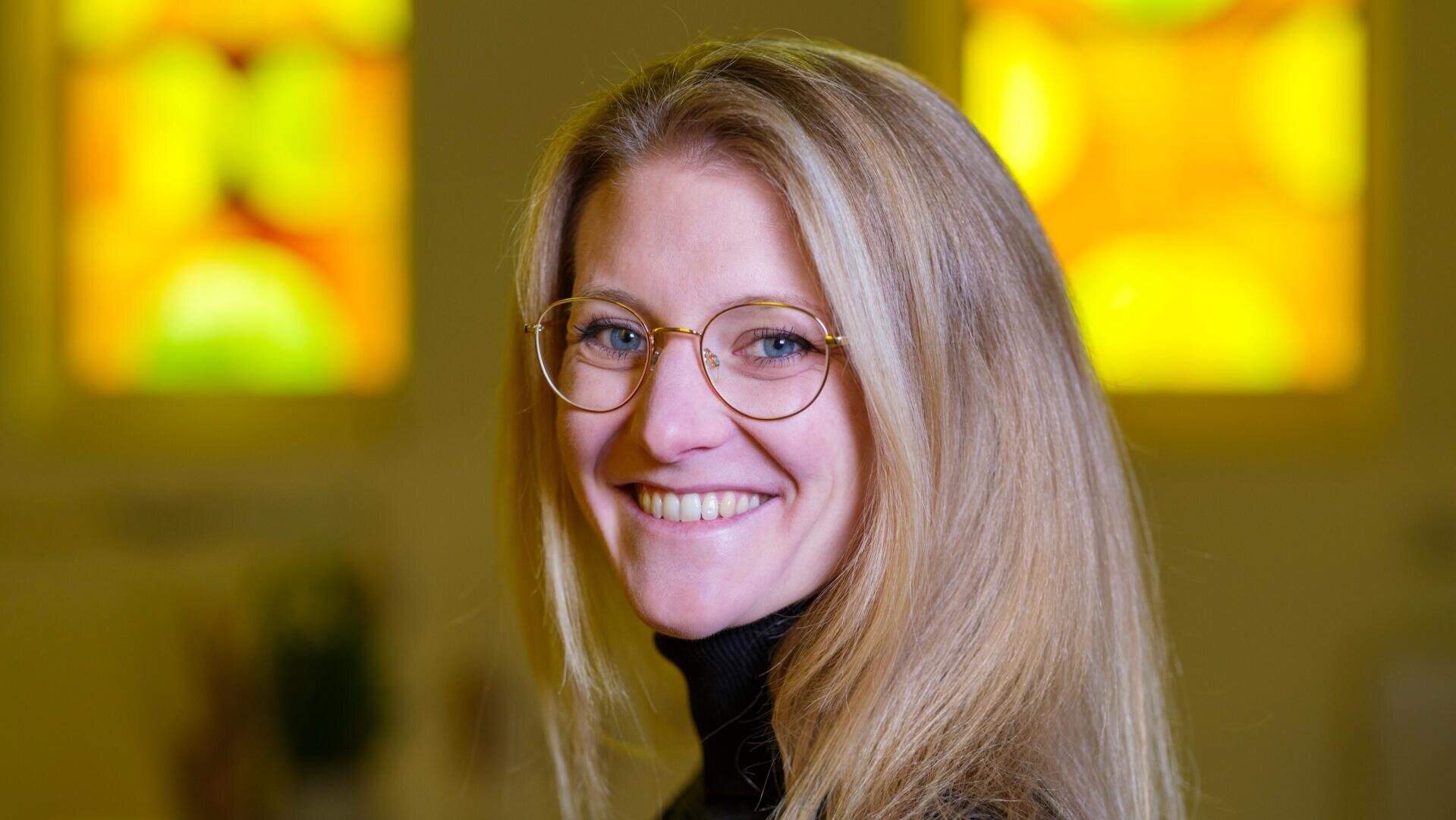 Eine Frau mit langen blonden Haaren und Brille lächelt in die Kamera. Sie trägt einen schwarzen Rollkragenpullover. Im unscharfen Hintergrund sind Buntglasfenster in Gelb- und Grüntönen zu sehen.