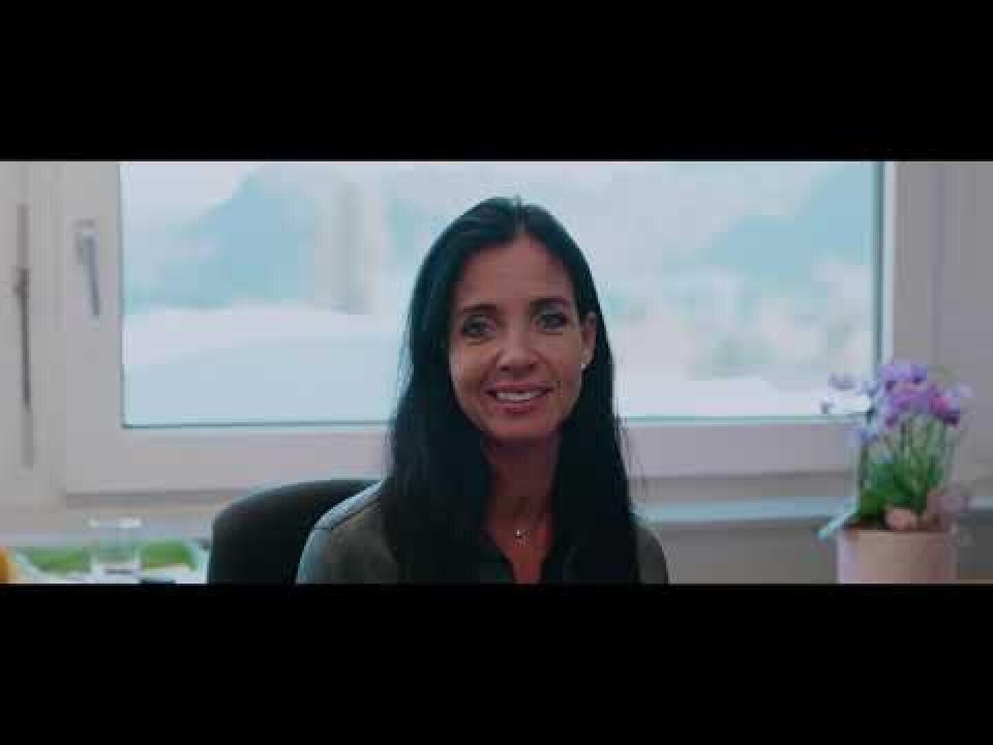 Eine Frau mit langen dunklen Haaren sitzt an einem Schreibtisch vor einem Fenster und lächelt in die Kamera. Auf der rechten Seite des Schreibtischs steht eine kleine Topfpflanze mit lila Blüten.