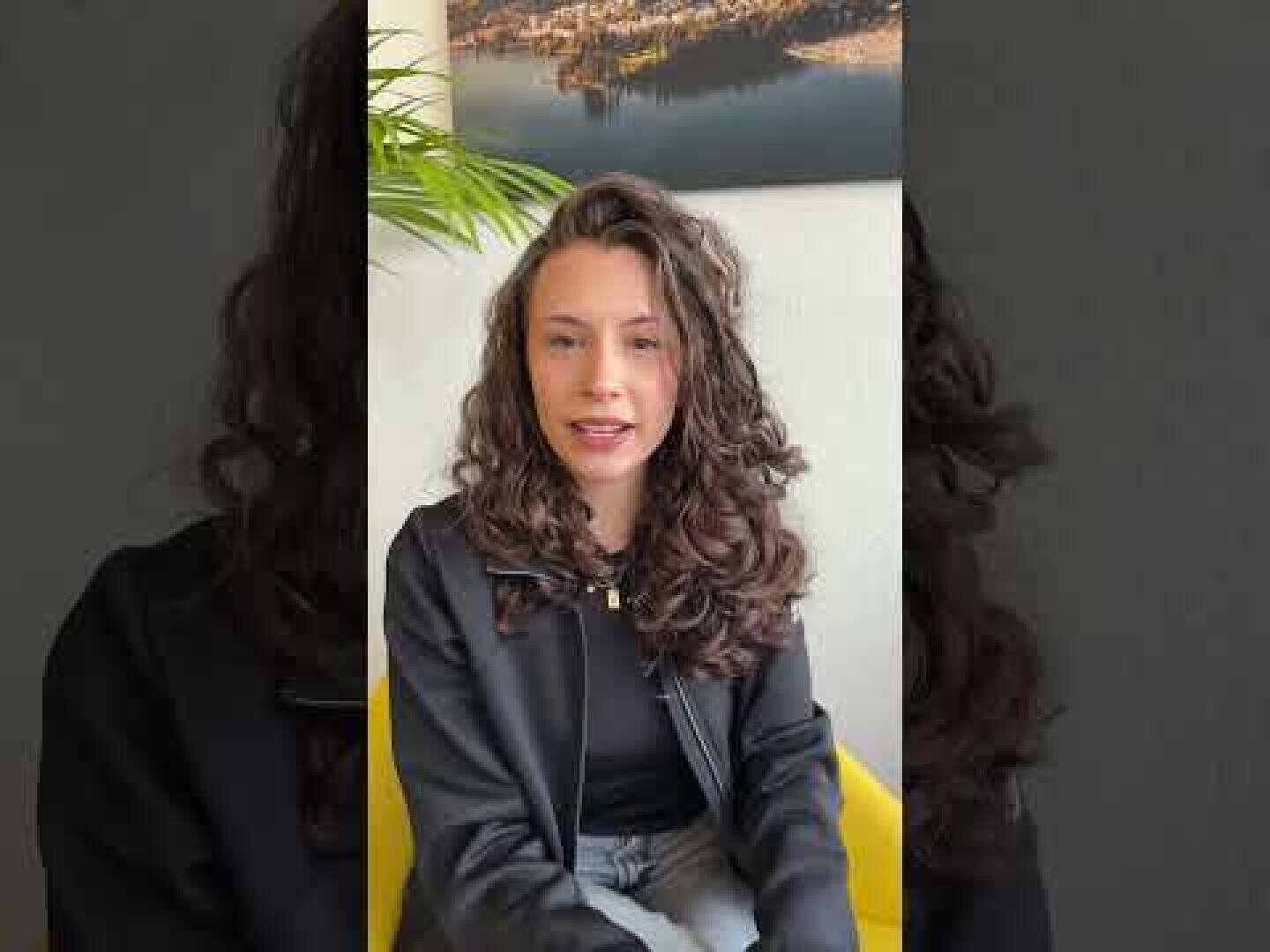 Eine Frau mit langen, lockigen braunen Haaren sitzt auf einem gelben Stuhl, trägt eine schwarze Jacke und eine graue Hose und hat einen neutralen Ausdruck. Im Hintergrund sind eine Grünpflanze und ein gerahmtes Kunstwerk zu sehen.