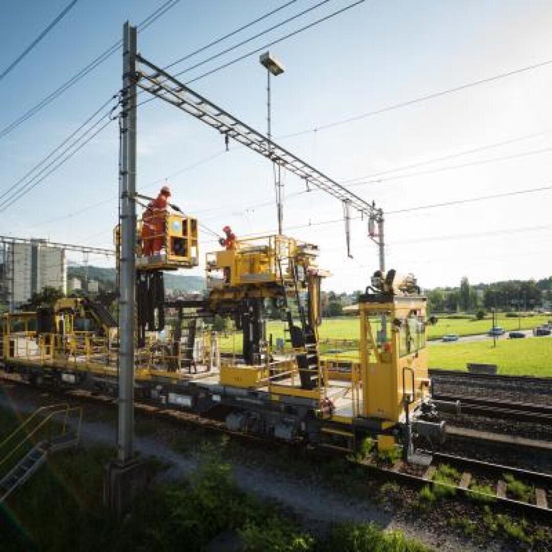 Arbeiter in orangefarbener Schutzkleidung bedienen ein gelbes Wartungsfahrzeug auf Eisenbahnschienen und benutzen einen Aufzug, um an die Oberleitungen zu gelangen. Im Hintergrund sind grüne Felder und Gebäude bei klarem Himmel zu sehen.