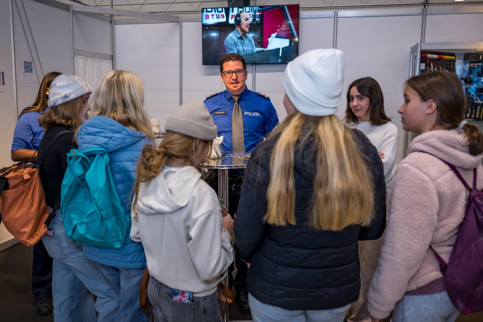 Ein Polizeibeamter in Uniform spricht mit einer Gruppe junger Leute in legerer Winterkleidung und mit Rucksäcken bei einer Indoor-Veranstaltung. Hinter dem Polizisten ist ein Fernsehbildschirm angebracht, auf dem eine Übertragung zu sehen ist.