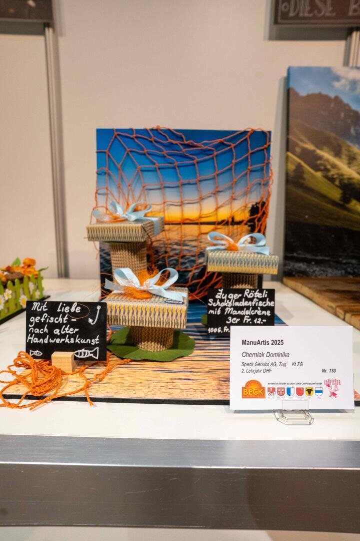 Drei dekorative Schachteln mit orangefarbenem Netz, die vor einem Foto einer Meereslandschaft ausgestellt sind, sind mit handgeschriebenen Kreidebeschriftungen auf Deutsch versehen. Vor dem Ausstellungsstück liegt eine Informationskarte zum Künstler.