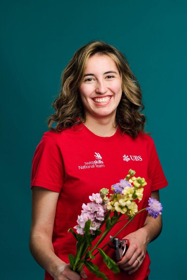 Vor einem blaugrünen Hintergrund steht eine lächelnde Person mit gewelltem Haar in einem roten SwissSkills-Nationalteam-Shirt und hält einen bunten Blumenstrauß sowie eine Gartenschere in der Hand.
