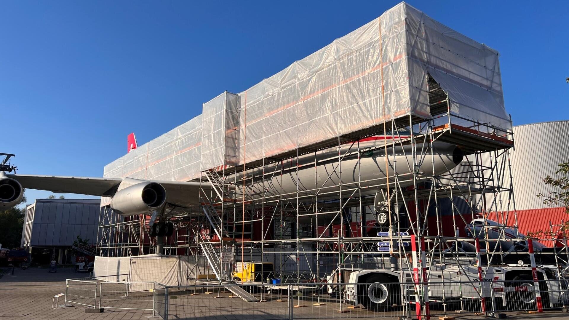 Ein großes Flugzeug ist von einem Gerüst umgeben und mit weißen Planen abgedeckt. Wahrscheinlich wird es gerade gewartet oder restauriert, während im Hintergrund der blaue Himmel zu sehen ist. Im Hintergrund sind Zäune und Baumaschinen zu sehen.