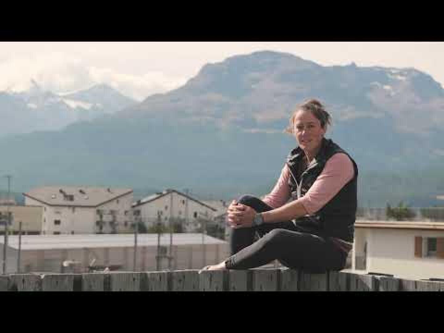 Eine Person sitzt auf einem Holzzaun und lächelt in die Kamera, während im Hintergrund Berge und Gebäude unter einem bewölkten Himmel zu sehen sind.