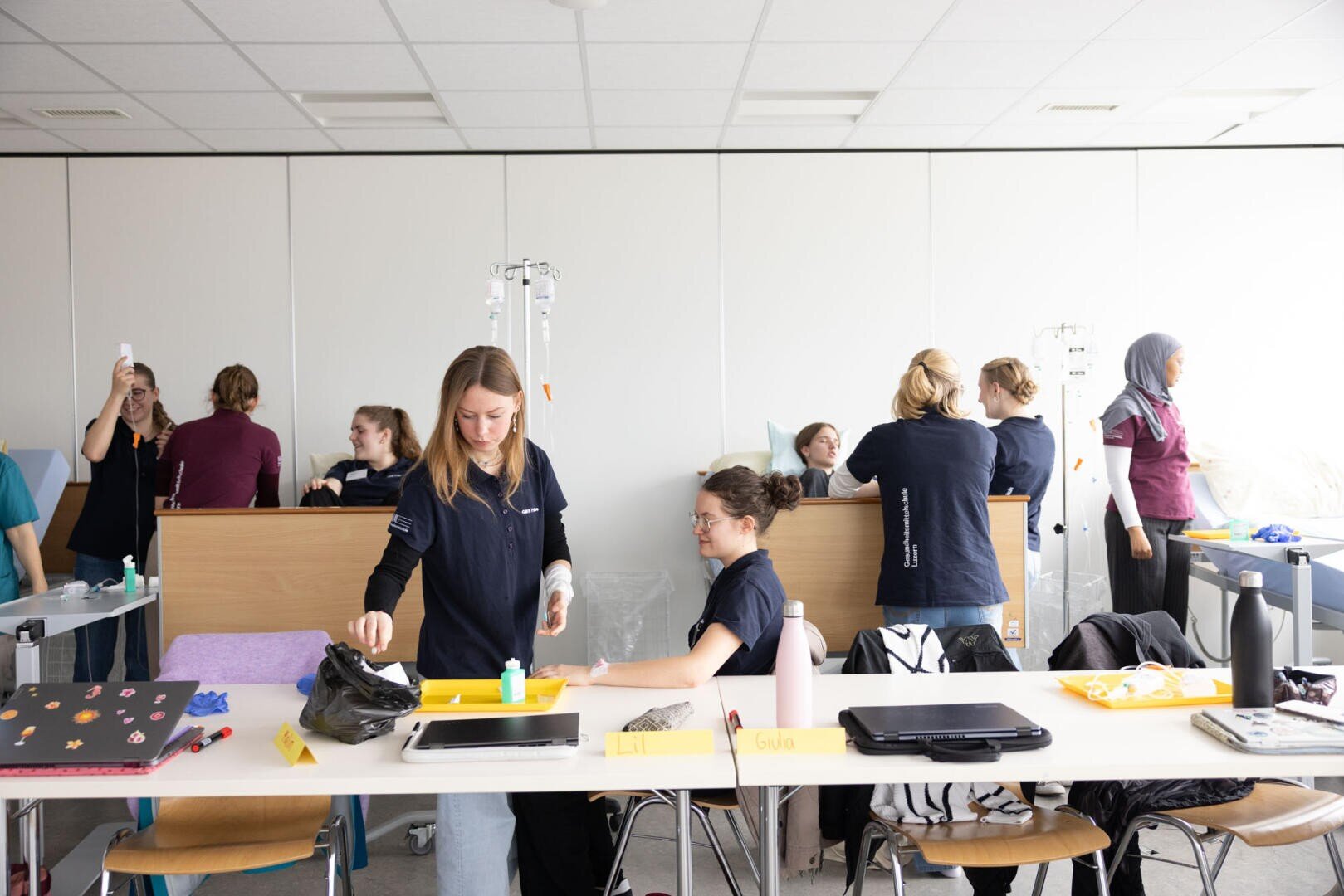 Eine Gruppe von Schülern übt in einem Klassenzimmer medizinische Verfahren. Einige arbeiten mit Infusionsständern, während andere an Tischen Material organisieren. Der Raum ist hell und geschäftig, auf den Tischen stehen Laptops und Wasserflaschen.