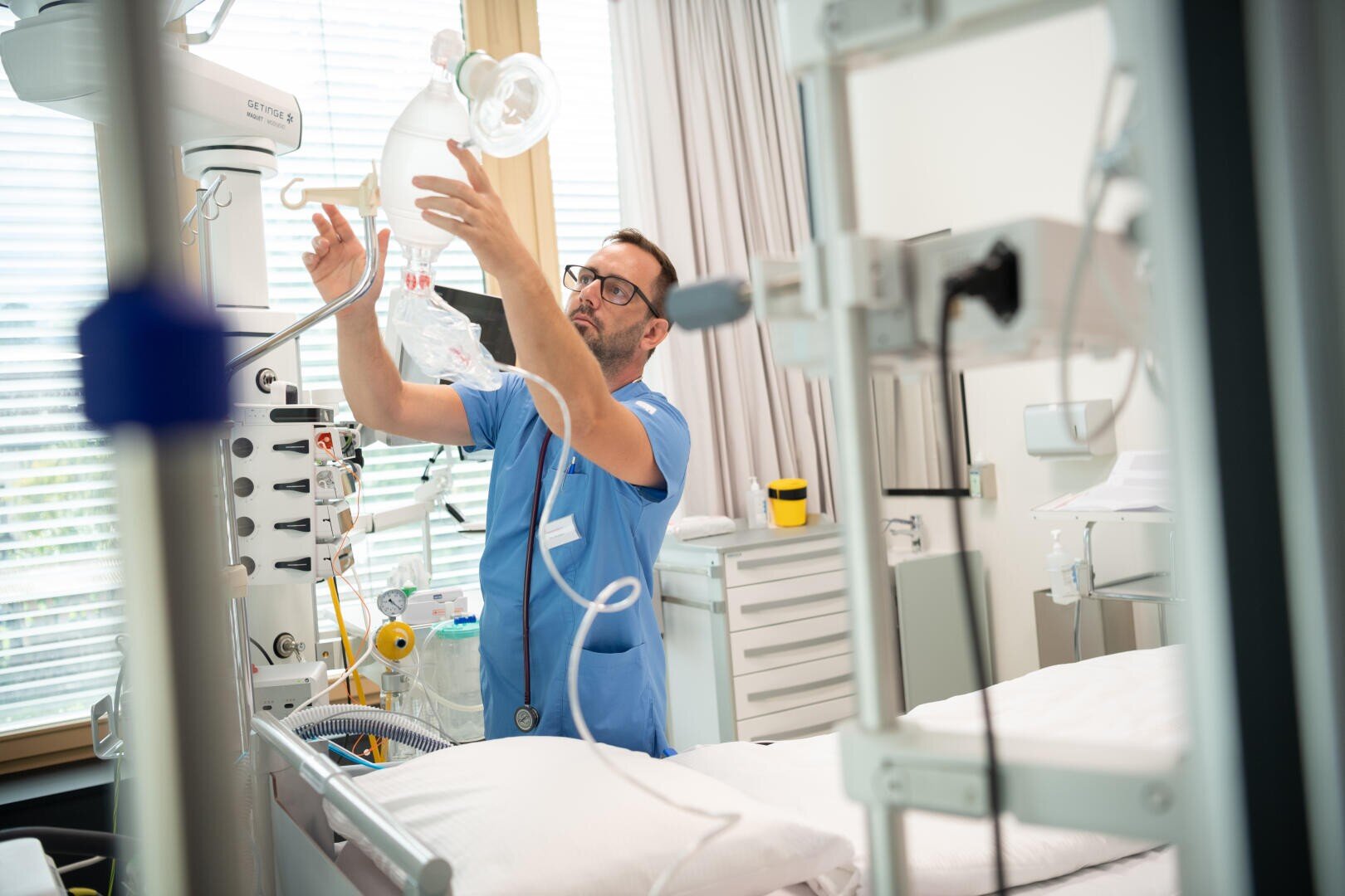 Ein Mitarbeiter des Gesundheitswesens in blauer OP-Kleidung justiert in einem hellen, modernen Krankenhauszimmer medizinische Geräte neben einem Krankenhausbett. Medizinische Geräte und Monitore umgeben das Bett, und das Sonnenlicht fällt durch die Jalousien.
