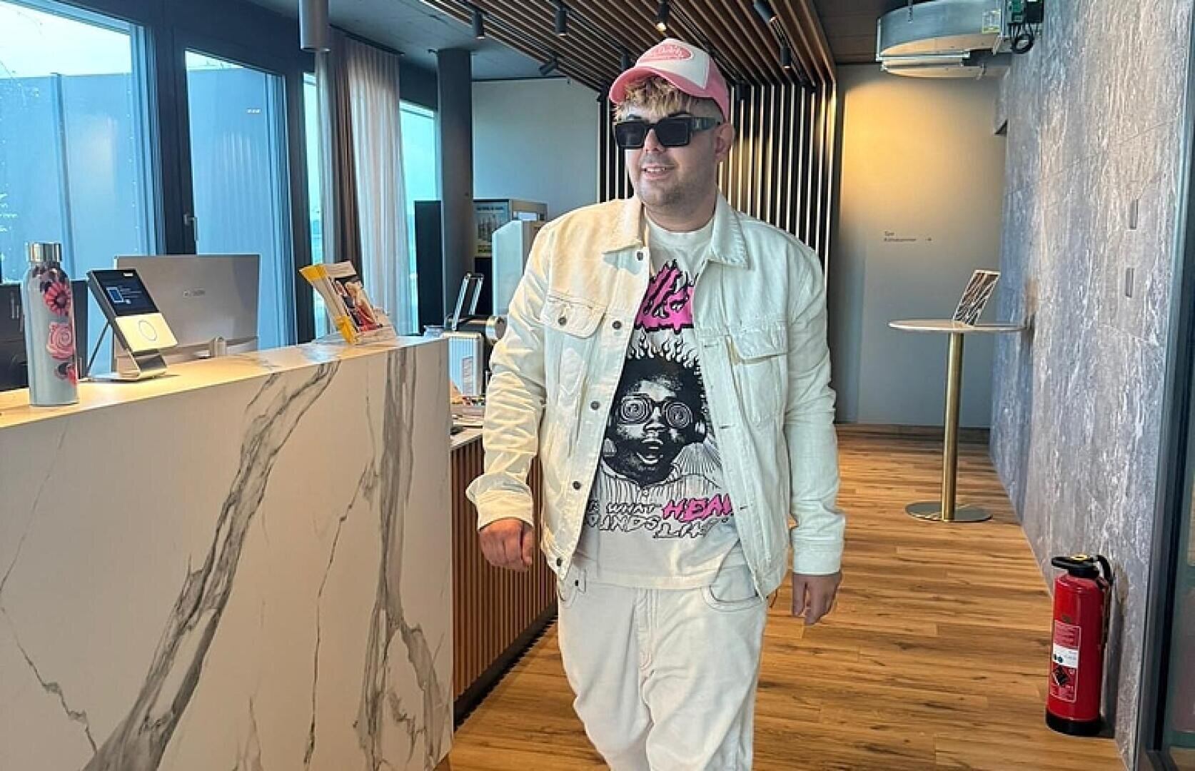 Eine Person, die eine weiße Jacke, ein grafisches T-Shirt, eine weiße Hose, eine rosa-weiße Kappe und eine Sonnenbrille trägt, steht in einem modernen Raum mit Holzfußboden und einem Empfangstresen aus Marmor. Rechts an der Wand ist ein Feuerlöscher zu sehen.