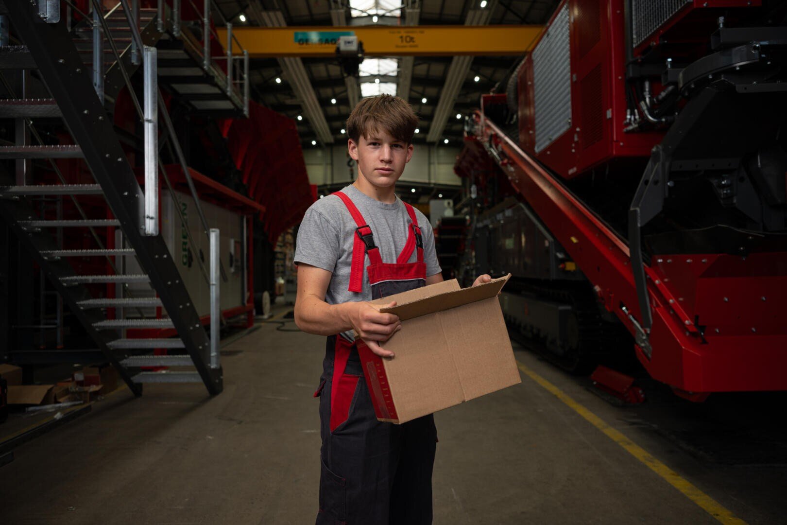 Ein junger Mann in Arbeitskleidung steht in einer Fabrik und hält einen offenen Karton in der Hand. Im Hintergrund sind große Industriemaschinen und Metalltreppen zu sehen.