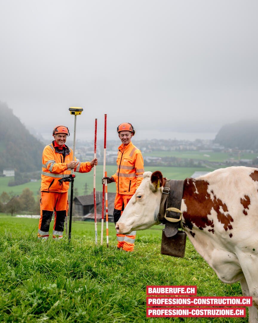 Zwei Vermessungsingenieure in orangefarbener Schutzkleidung stehen in einem grasbewachsenen Feld mit Messgeräten und lächeln in die Kamera. Im Vordergrund grast eine Kuh. Im nebligen Hintergrund sind Berge und ein See zu sehen. Der Werbetext befindet sich am unteren Rand.
