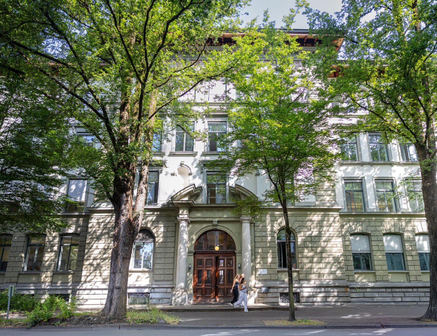 Ein großes, helles Steingebäude mit hohen Fenstern wird teilweise von grünen Bäumen beschattet. Zwei Personen gehen am Eingang vorbei, der aus Säulen und einer Holztür besteht. Das Sonnenlicht fällt durch die Äste.
