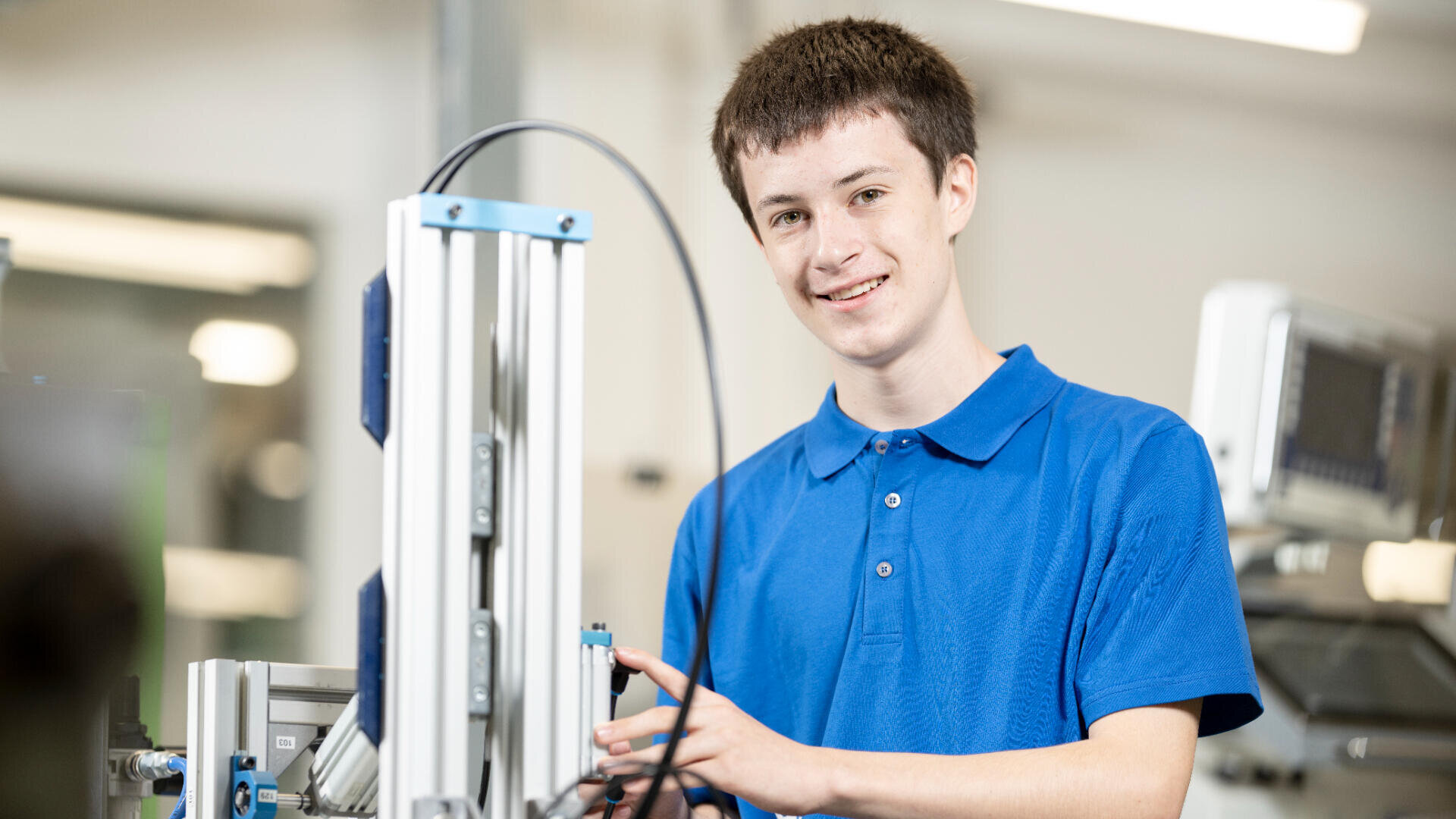 Ein junger Mensch in einem blauen Polohemd lächelt, während er in einem modernen, hell erleuchteten technischen oder ingenieurwissenschaftlichen Labor mit Geräten arbeitet.
