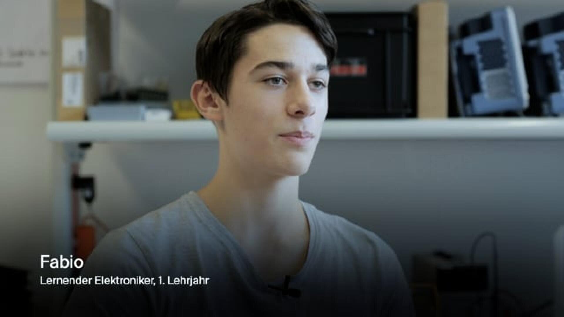 Ein junger Mann mit kurzen dunklen Haaren sitzt in einem Haus und blickt leicht nach links. Der Text auf dem Bild lautet: Fabio, Lernender Elektroniker, 1. Lehrjahr. Im Hintergrund sind Regale mit Elektronik und Geräten zu sehen.