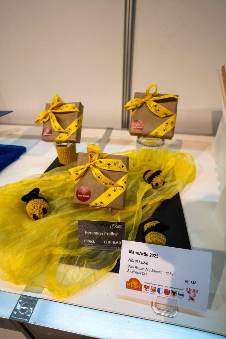 Geschenkboxen mit gelben Schleifen stehen auf einem gelben Tuch mit gehäkelten Bienen. Auf einer Karte sind die Produktdetails und Preise für „Bärnker Praliné“ auf einer Messe aufgeführt.