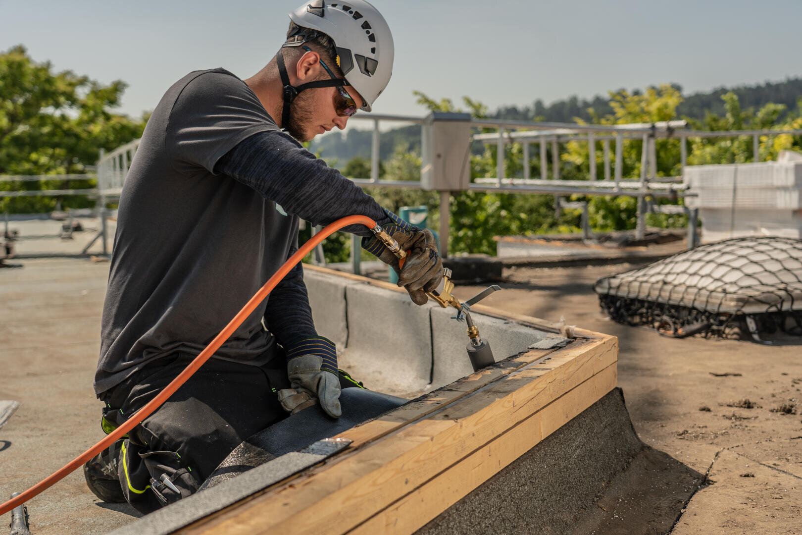 Ein Bauarbeiter, der einen Helm und Handschuhe trägt, arbeitet an einem sonnigen Tag im Freien mit einem Brenner an Dachmaterial, umgeben von Sicherheitsausrüstung und Grünpflanzen.