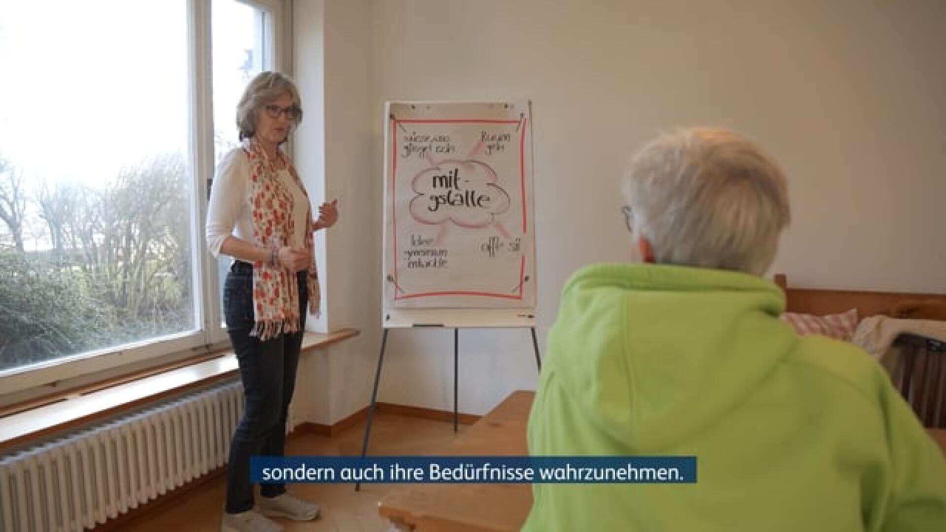 Eine Frau steht neben einem Flipchart und erklärt einer sitzenden Person etwas; in den deutschen Untertiteln heißt es: „sondern auch ihre Bedürfnisse wahrzunehmen…“.
