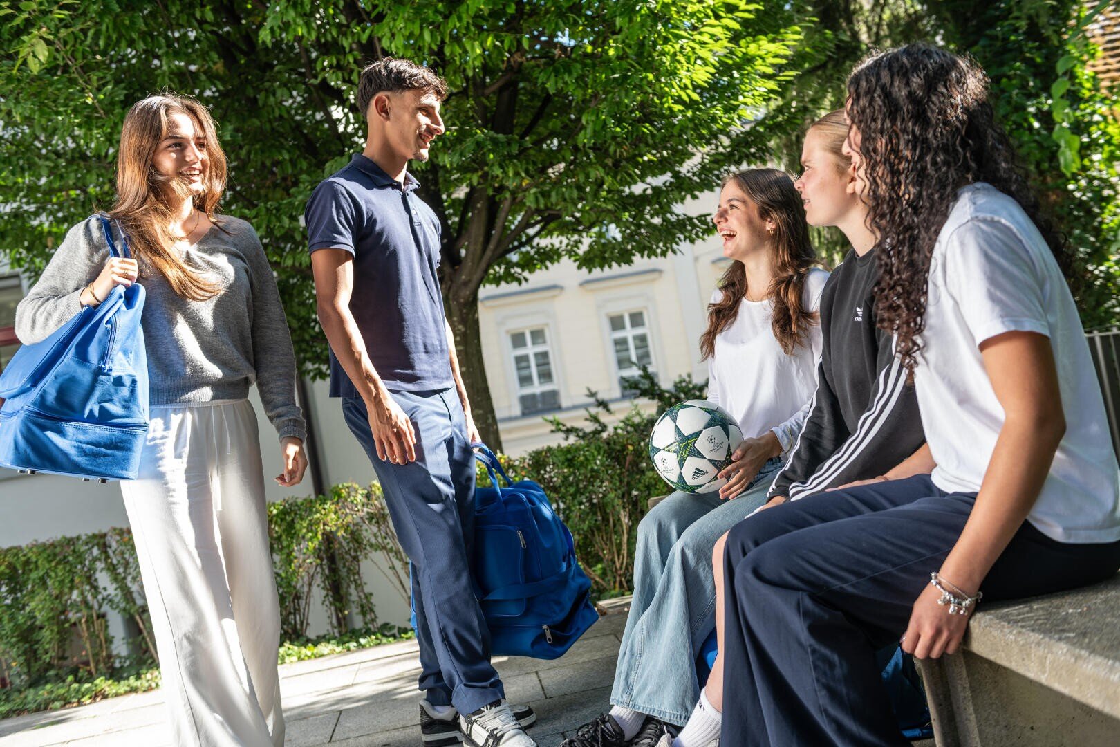 Eine Gruppe von fünf lächelnden Teenagern, einige mit Sporttaschen und einem Fußball in der Hand, versammelt sich an einem sonnigen Tag im Freien, redet und lacht in der Nähe von grünen Bäumen und einem Gebäude im Hintergrund.