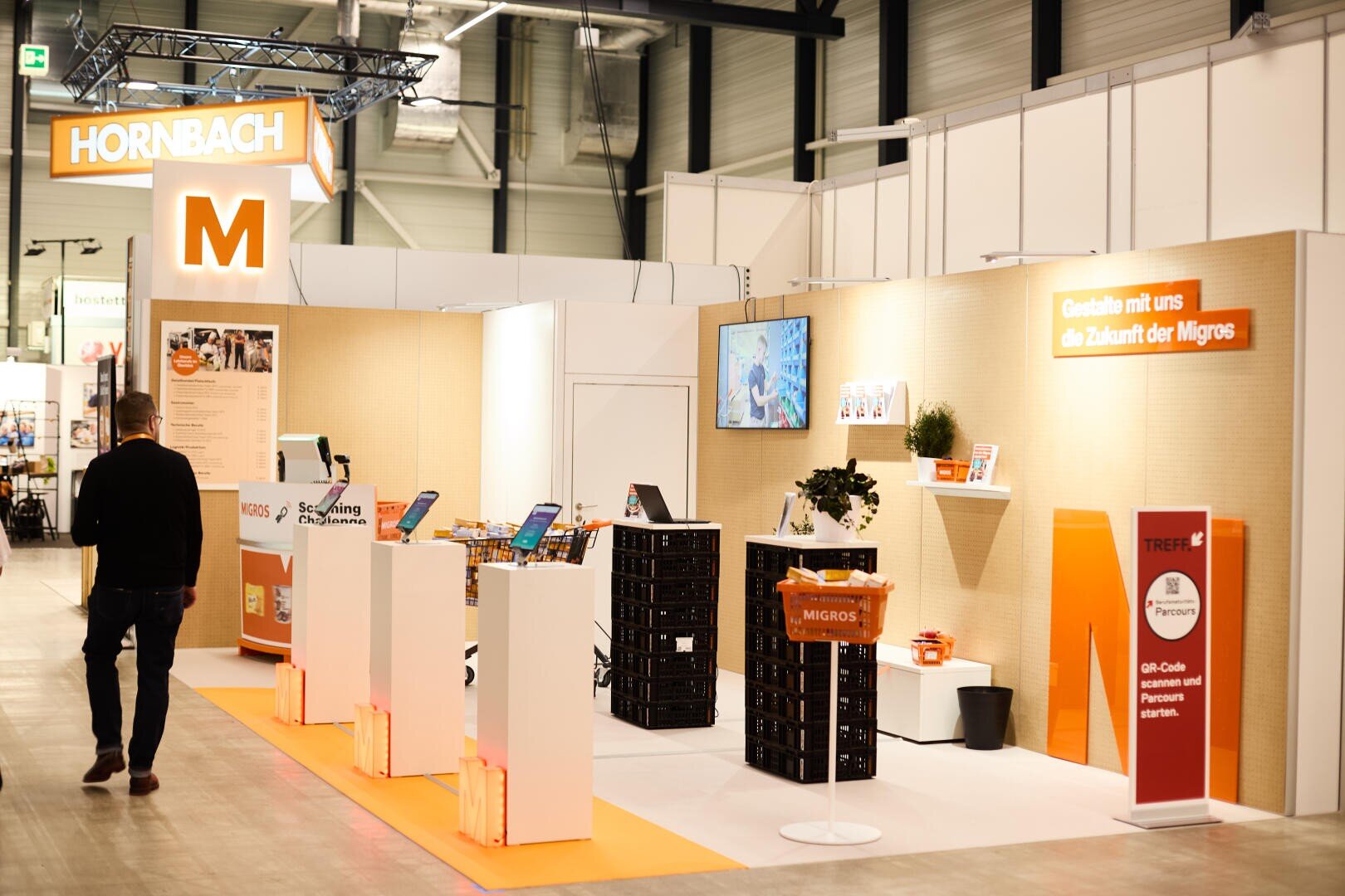 Ein Mann geht an einem hellen Messestand mit Hornbach- und Migros-Branding vorbei, der mit Informationsschaltern, digitalen Bildschirmen und Werbedisplays in einer modernen Veranstaltungshalle ausgestattet ist.