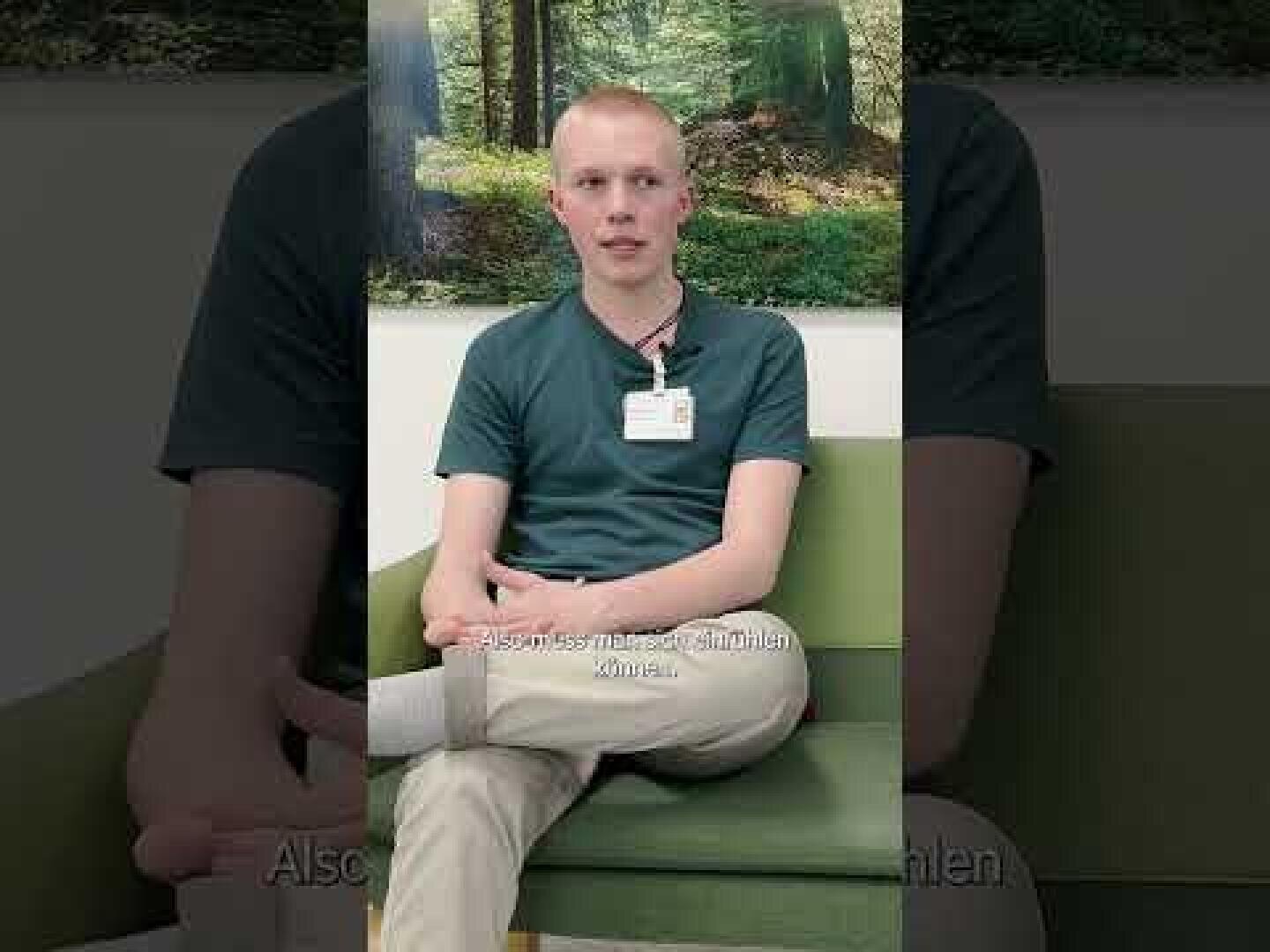 Eine Person mit kurzen blonden Haaren, grünem Hemd und heller Hose sitzt mit gekreuzten Beinen auf einer grünen Couch. Sie trägt ein Namensschild und spricht, hinter ihr hängt ein Waldfoto an der Wand.