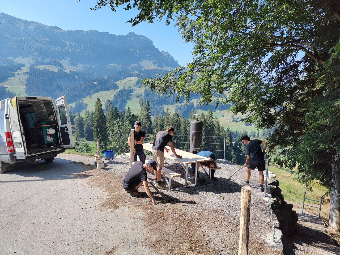 Vier Personen reinigen und decken einen Picknicktisch in der Nähe eines weißen Lieferwagens, im Hintergrund sind Berge, Bäume und ein klarer Himmel zu sehen. Eine Person wischt den Tisch ab, während die anderen den Boden fegen und Unrat aufsammeln.