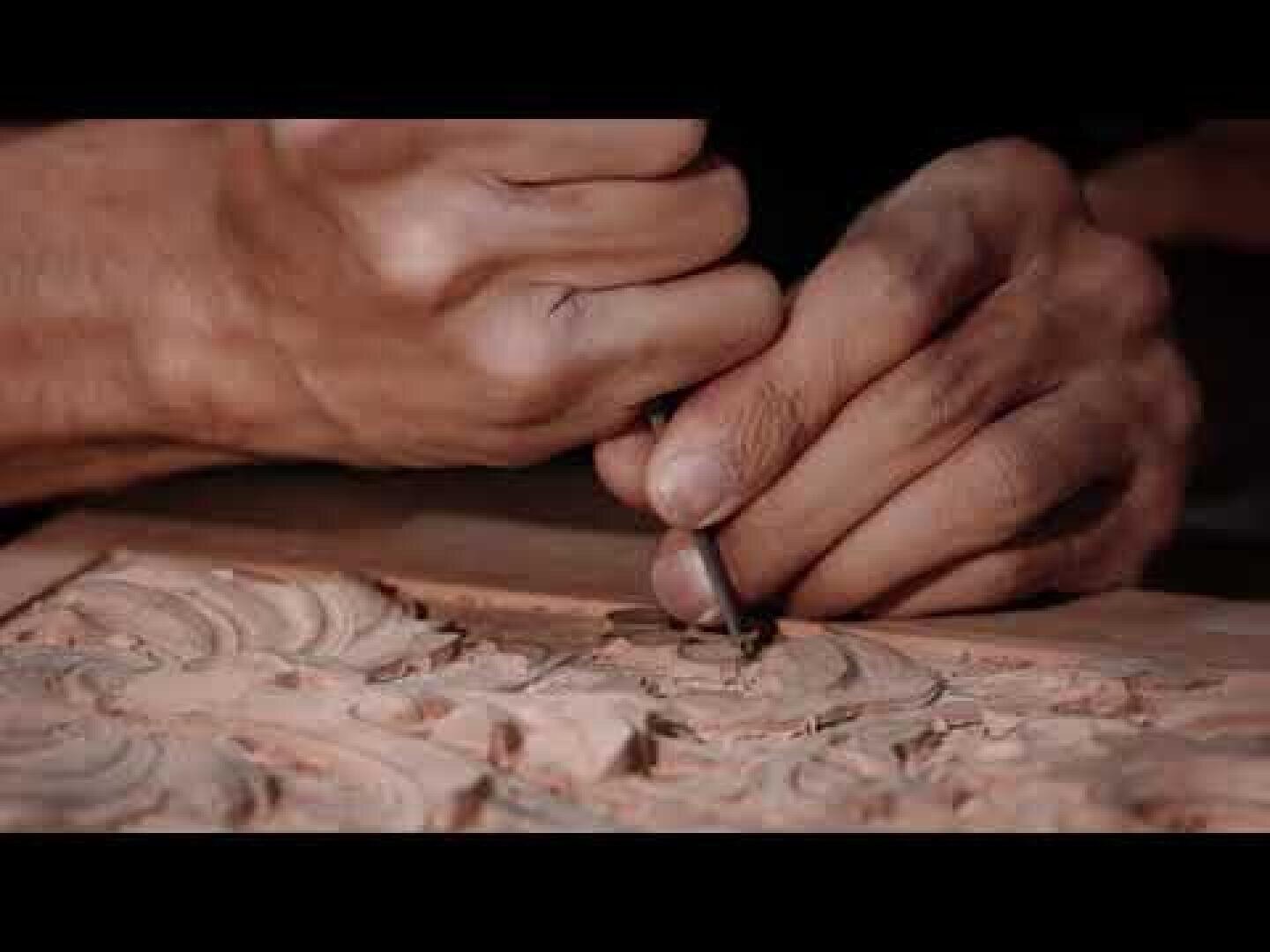 Nahaufnahme von Händen, die mit einem Schnitzwerkzeug komplizierte Muster in ein Stück Holz schnitzen, die detaillierte Handwerkskunst und feine Holzbearbeitung zeigen.